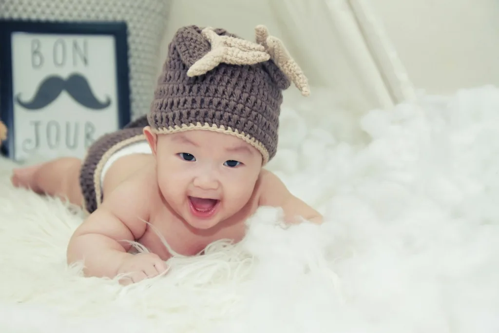 A joyful baby wearing a brown knitted hat lies on a soft surface, radiating happiness.
